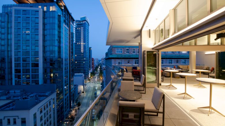 A modern balcony with tables and chairs overlooks a cityscape filled with tall buildings and lit streets at dusk, creating an urban ambiance.