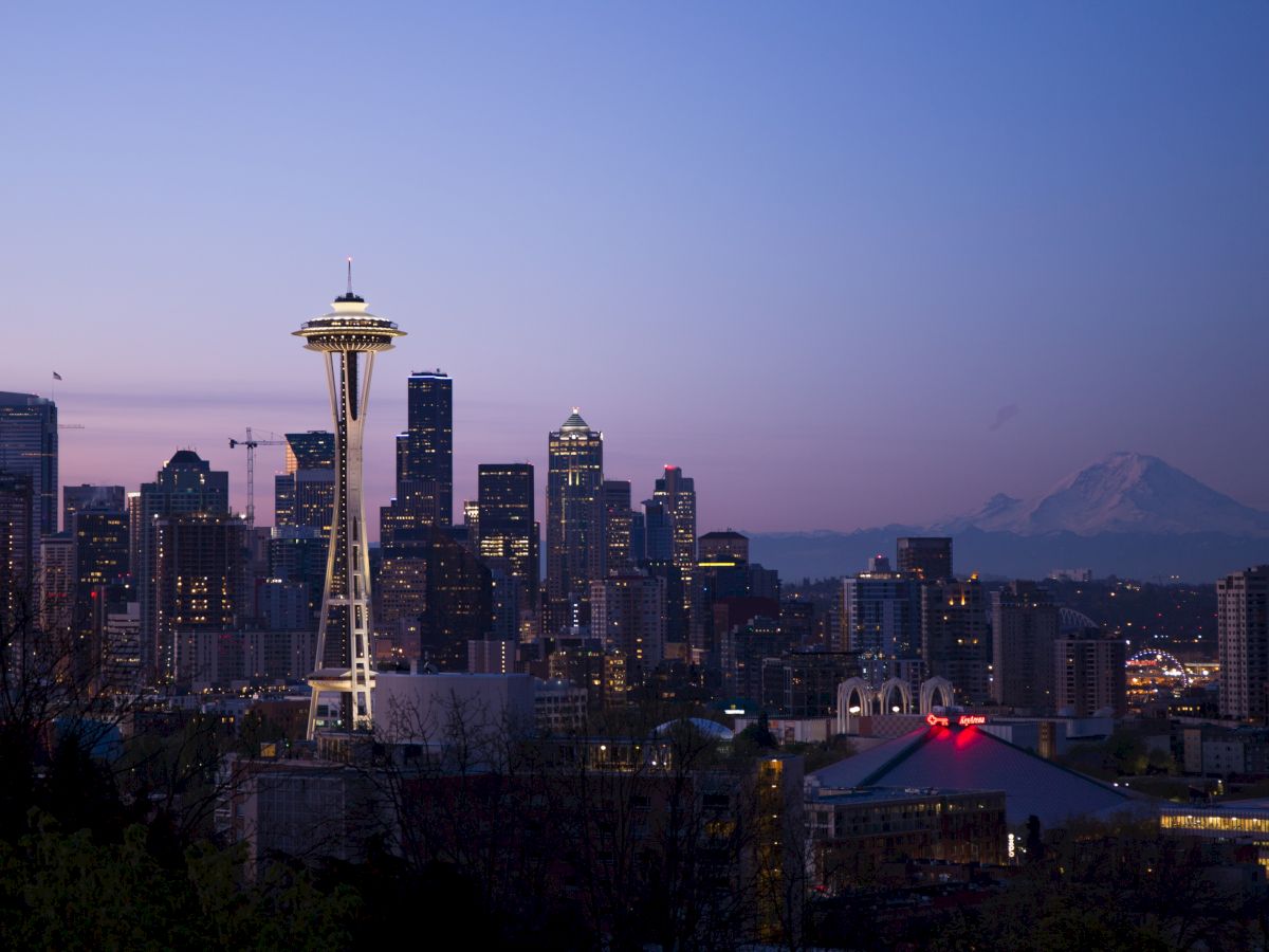 The image shows a twilight view of the Seattle skyline, featuring the Space Needle and Mount Rainier in the background, with the city lights beginning to illuminate.