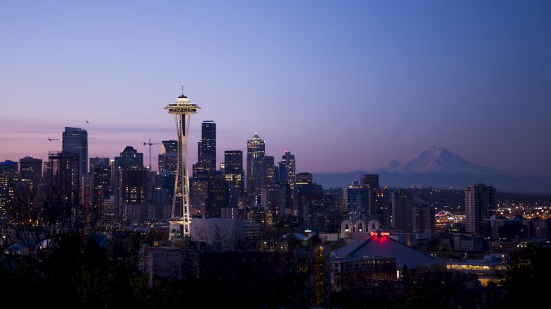 The image shows a twilight view of the Seattle skyline, featuring the Space Needle and Mount Rainier in the background, with the city lights beginning to illuminate.