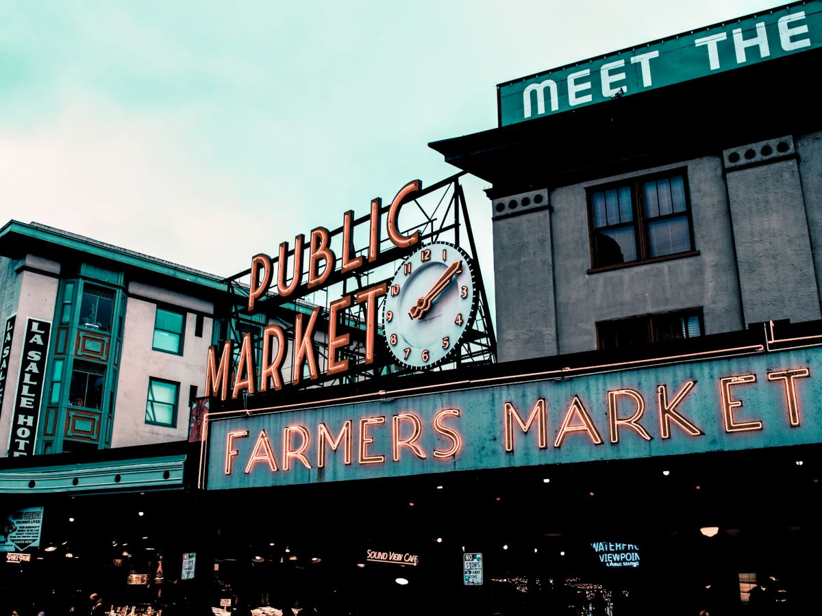The image shows the iconic "Public Market" and "Farmers Market" signs, surrounded by buildings and a clock, evoking a classic market scene.