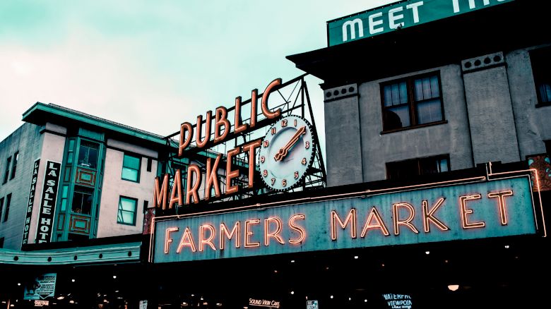 The image shows the iconic "Public Market" and "Farmers Market" signs, surrounded by buildings and a clock, evoking a classic market scene.