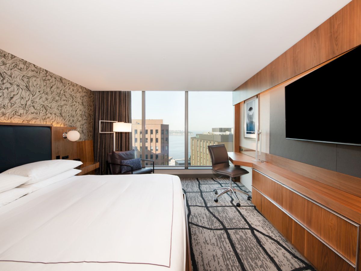 A modern hotel room with a large bed, artwork on the wall, a desk, chair, and a big TV facing the bed. The window offers a city view.