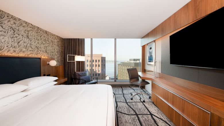 A modern hotel room with a large bed, artwork on the wall, a desk, chair, and a big TV facing the bed. The window offers a city view.
