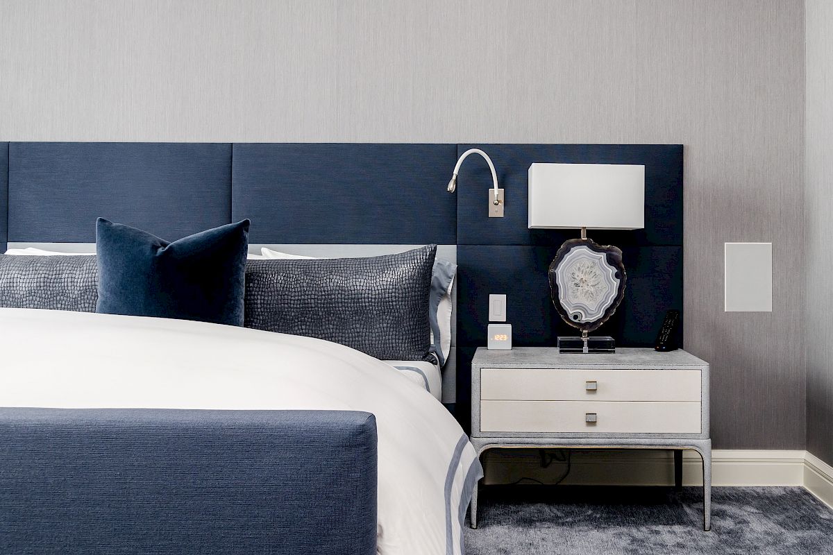 A modern bedroom with a blue headboard, bed with white and blue linens, a white nightstand with a lamp, and a gray carpet.