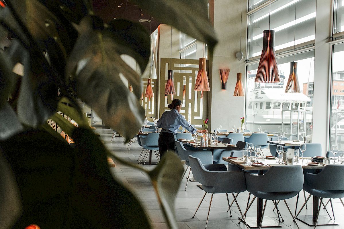 A restaurant interior with modern decor, including pendant lights and blue chairs, as an employee arranges tables near large windows.