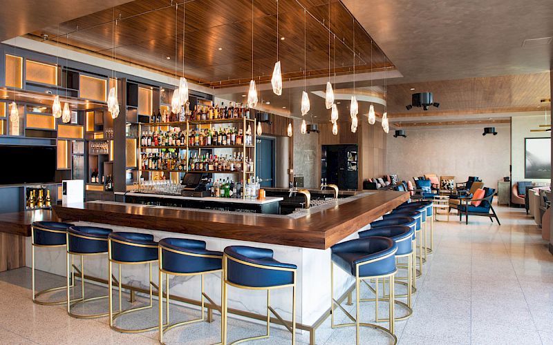A stylish bar with blue cushioned barstools, a variety of drinks behind the counter, pendant lights, and a seating area in the background.