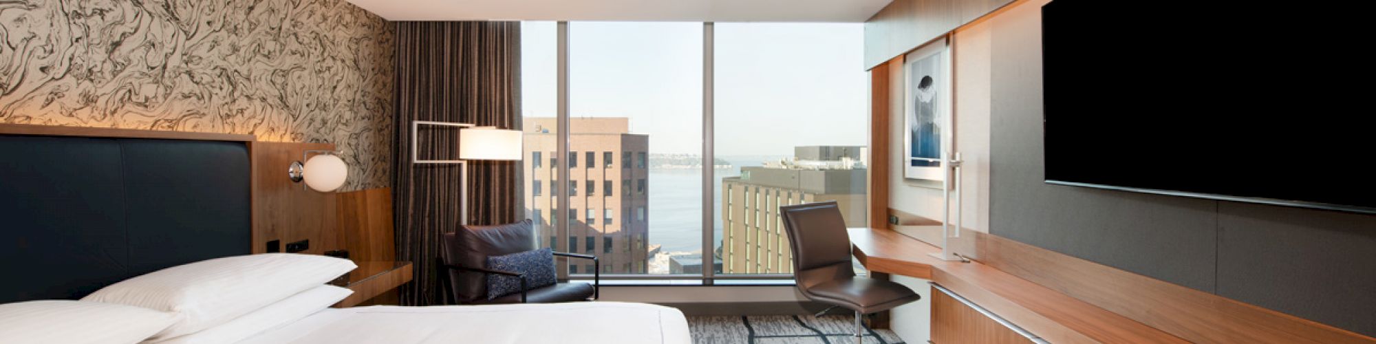 A modern hotel room featuring a king-sized bed, a large TV, a desk, a chair, and a window with a cityscape view in the background.