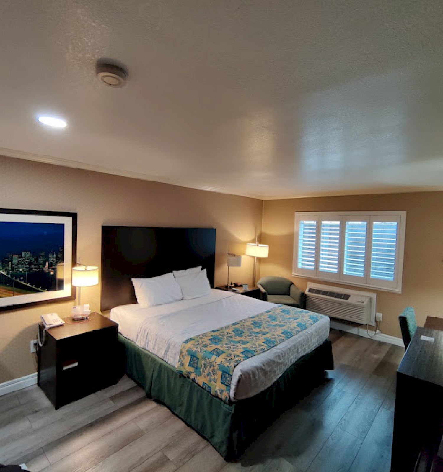 A hotel room with a bed, lamps, a window with shutters, a desk, TV, and a chair. There is also a framed picture on the wall and wooden flooring.