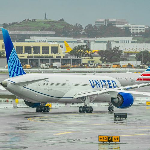 An airport scene with two commercial airplanes, one from United Airlines and the other with a striped tail, parked on the wet tarmac on a rainy day.