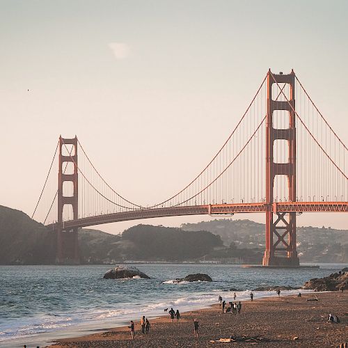 A scenic view of a beach with people, and a large suspension bridge in the background with hills and water.