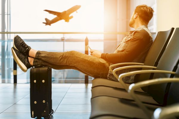 A person sits in an airport lounge with feet on a suitcase, watching an airplane take off in the distance.