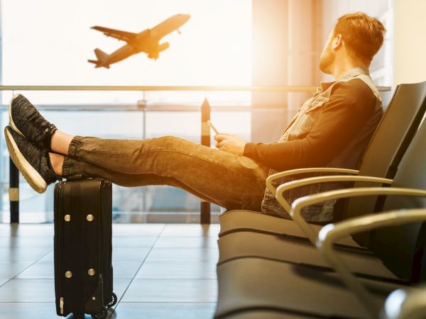 A person sits in an airport lounge with feet on a suitcase, watching an airplane take off in the distance.